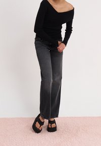 Personne portant un pull noir à épaules dénudées, un jean noir délavé et des sandales à plateforme noires, debout sur un tapis rose clair.