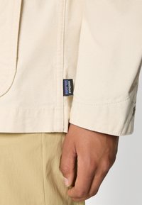 Close-up van een hand naast een crème-kleurige jasmanchet met een klein Patagonia-label, gecombineerd met beige broeken.