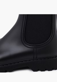 Bota negra impermeable hasta el tobillo con un acabado de goma suave, paneles laterales elásticos y una suela estriada para mayor tracción.