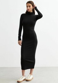Robe à col roulé noir à manches longues, coupe cintr�ée, en tissu doux, atteignant le mi-mollet, associée à des baskets blanches et beiges.