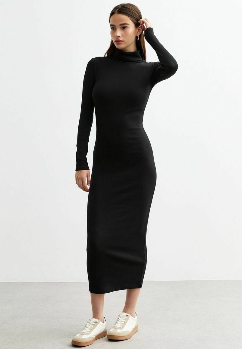 Robe à col roulé noir à manches longues, coupe cintrée, en tissu doux, atteignant le mi-mollet, associée à des baskets blanches et beiges.