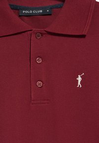 Polo color burdeos con tejido texturizado, que presenta tres botones, un cuello de solapa y un pequeño logo de un golfista en blanco en el lado izquierdo del pecho.