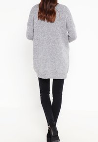 Femme aux longs cheveux bruns portant un pull en tricot gris surdimensionné, un pantalon skinny noir et des bottines noires à talons, se tenant de dos.