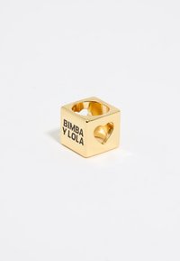 Gold-toned cubic ring with a hollow center, features a heart-shaped cutout, and displays "BIMBA Y LOLA" text on one side.