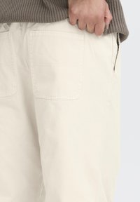 Light beige trousers with a smooth texture, featuring two back pockets and a side pocket, paired with a knitted, neutral-toned sweater.