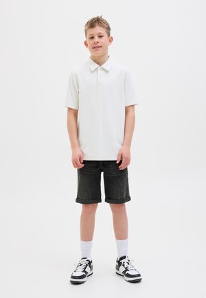 Băiat stând cu fața înainte, purtând un tricou polo alb, pantaloni scurți negri suflecați, șosete albe și adidași alb-negru pe un fundal alb.