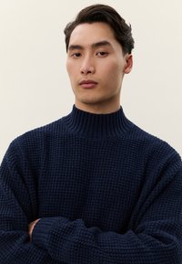 Mlad moški s temnimi lasmi, oblečen v teksturiran temno moder pleten pulover z visokim ovratnikom, gleda neposredno naprej z rokami prekrižanimi na prsih.