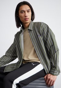 Camisa de botones a rayas verdes sobre una camisa beige, combinada con pantalones negros con franjas laterales blancas y el logo de Puma. Textura de tela suave.