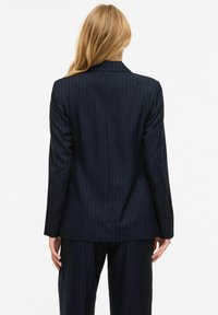 Femme blonde debout, dos tourné vers la caméra, portant un blazer à fines rayures bleu marine et un pantalon assorti sur fond blanc.