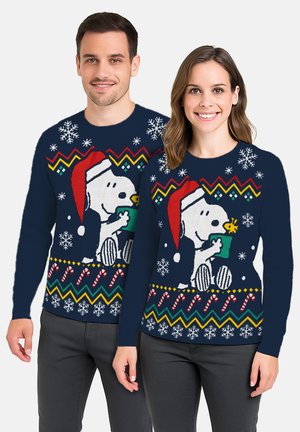 Mann und Frau tragen passende dunkelblaue Weihnachtspullover mit Snoopy in einem Weihnachtsmannhut und weihnachtlichen Mustern, darunter Schneeflocken und Zuckerstangen.