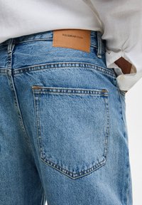 Jeans i ljusblå denim, med en läderetikett som har texten "PULL&BEAR DENIM", femficksdesign och synliga sömdetaljer.