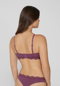 Femme aux longs cheveux bruns vue de dos, portant un ensemble de lingerie violet avec dentelle assortie, contre un fond clair.