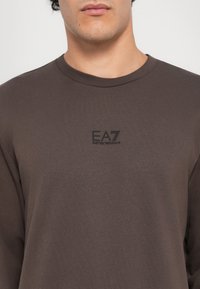 Maglietta marrone a maniche lunghe con scollatura rotonda e un piccolo logo EA7 nero. Il tessuto appare liscio con una texture morbida.
