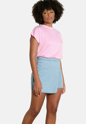 Woman with curly hair wearing a pink short-sleeve top and light blue denim wrap skirt standing against a plain background.