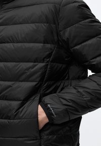 Main dans la poche d'une veste noire matelassée avec des coutures horizontales et le logo Jack & Jones sur le poignet.