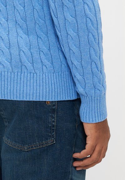 Φωτεινό γαλάζιο πουλόβερ με πλέξη καλτσών, ριμπ τελείωμα και μανσέτες, συνδυασμένο με σκουρόχρωμο τζιν. Κοντινή λήψη του μανικιού και του χεριού.