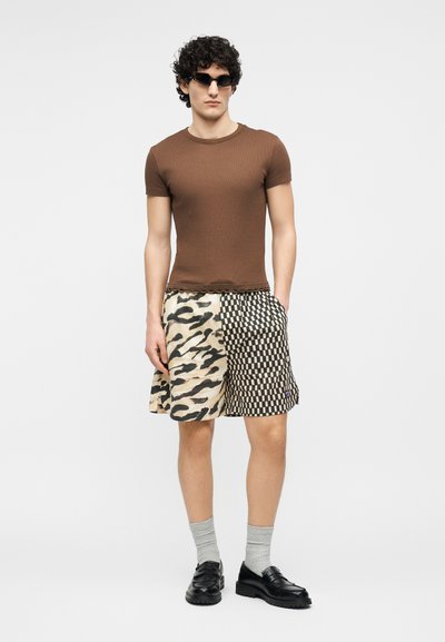 Homme portant un t-shirt marron ajusté, des lunettes de soleil noires, un short à motif partagé avec imprimé animalier et damier, des chaussettes grises et des mocassins noirs.