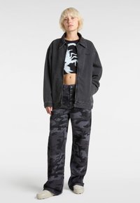 Giacca di jeans nera oversize sopra una maglietta nera graphic cropped, abbinata a pantaloni larghi con motivo camouflage e scarpe di colore chiaro.