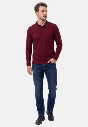 Mand iført vinrød langærmet poloshirt, blå jeans og sorte sko, stående med den ene hånd i lommen og kiggede mod højre.