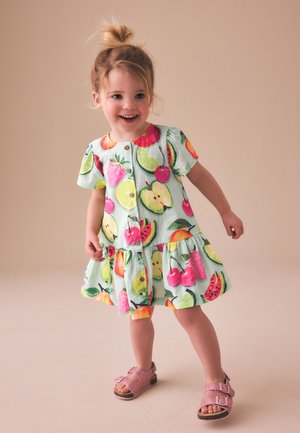 Lille pige der smiler og står op, iført en kortærmet kjole med farverige frugtprint og pink sandaler mod en neutral baggrund.