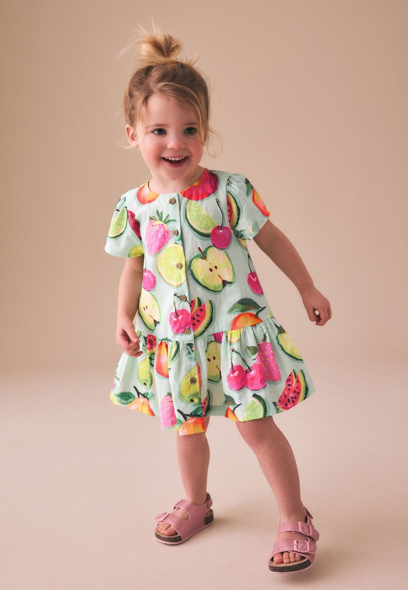 Gyermeklány mosolyogva áll, rövid ujjú ruha színes gyümölcsmintákkal és rózsaszín szandálok, semleges háttér előtt.