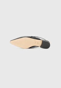 Black patent leather pointed-toe shoe with a beige sole and black heel. Features a thin strap and smooth texture. Model name underfoot.