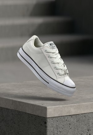 Baskets Converse All Star blanches basses suspendues au-dessus d'un rebord en béton avec des escaliers flous en arrière-plan.