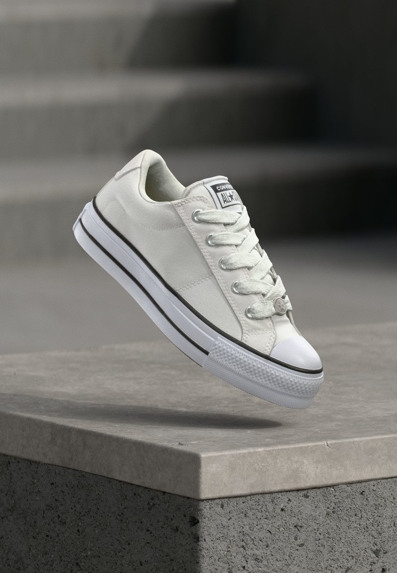 Λευκό Converse All Star sneaker χαμηλού προφίλ, αιωρούμενο πάνω από μια τσιμεντένια άκρη με θολά σκαλοπάτια στο φόντο.