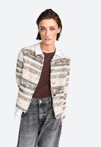 Giacca testurizzata con una combinazione di strisce crema, marrone e grigio, con una camicia bianca con colletto sotto e pantaloni di jeans grigi.