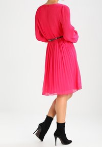 Růžové plisované šaty s dlouhými rukávy a černým cvočkovaným opaskem. Mají volný siluetu, hladkou texturu a délku do kolen. Černé vysoké kozačky.