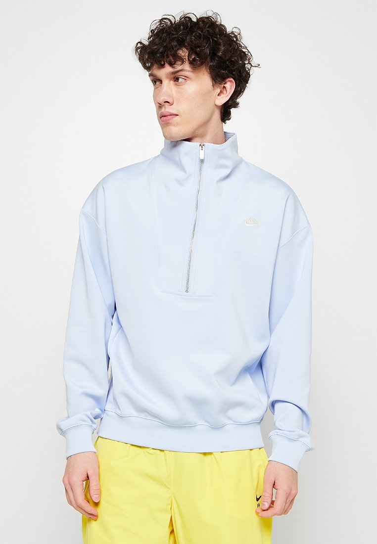 Ljusblå fleece-tröja med halv dragkedja, elastiska ärmslut och en diskret Nike-logotyp på bröstet. Slät textur och afslappnad passform.