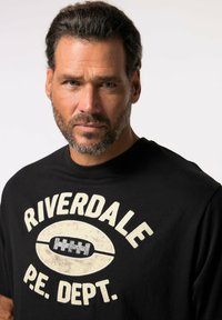 Camiseta de algodón negra con el texto "RIVERDALE P.E. DEPT." en color crema, con un gráfico de fútbol, diseño de cuello redondo y detalle de impresión texturizada.