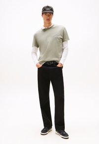 Graugrünes T-Shirt über einem weißen Langarmshirt; schwarze Hose; schwarze Kappe mit Logo; schwarze Schuhe mit weißen Sohlen; entspannter Schnitt.