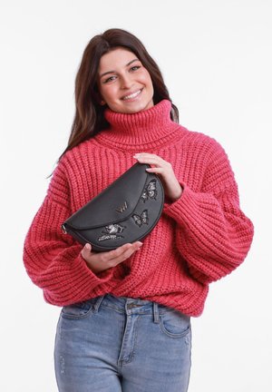 Černá kožená kabelka s vyšitými motýly, zakřivený tvar, stříbrný logový akcent, držena osobou v růžovém svetru s hrubým pletením.