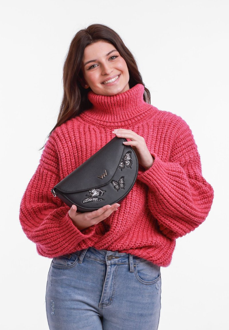 Černá kožená kabelka s vyšitými motýly, zakřivený tvar, stříbrný logový akcent, držena osobou v růžovém svetru s hrubým pletením.