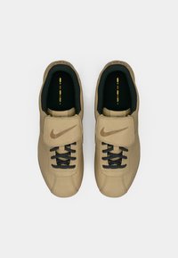 Adidași Nike bej cu un superior din piele netedă, șireturi negre și o bandă Velcro. Talpă din cauciuc texturat și interior negru căptușit.