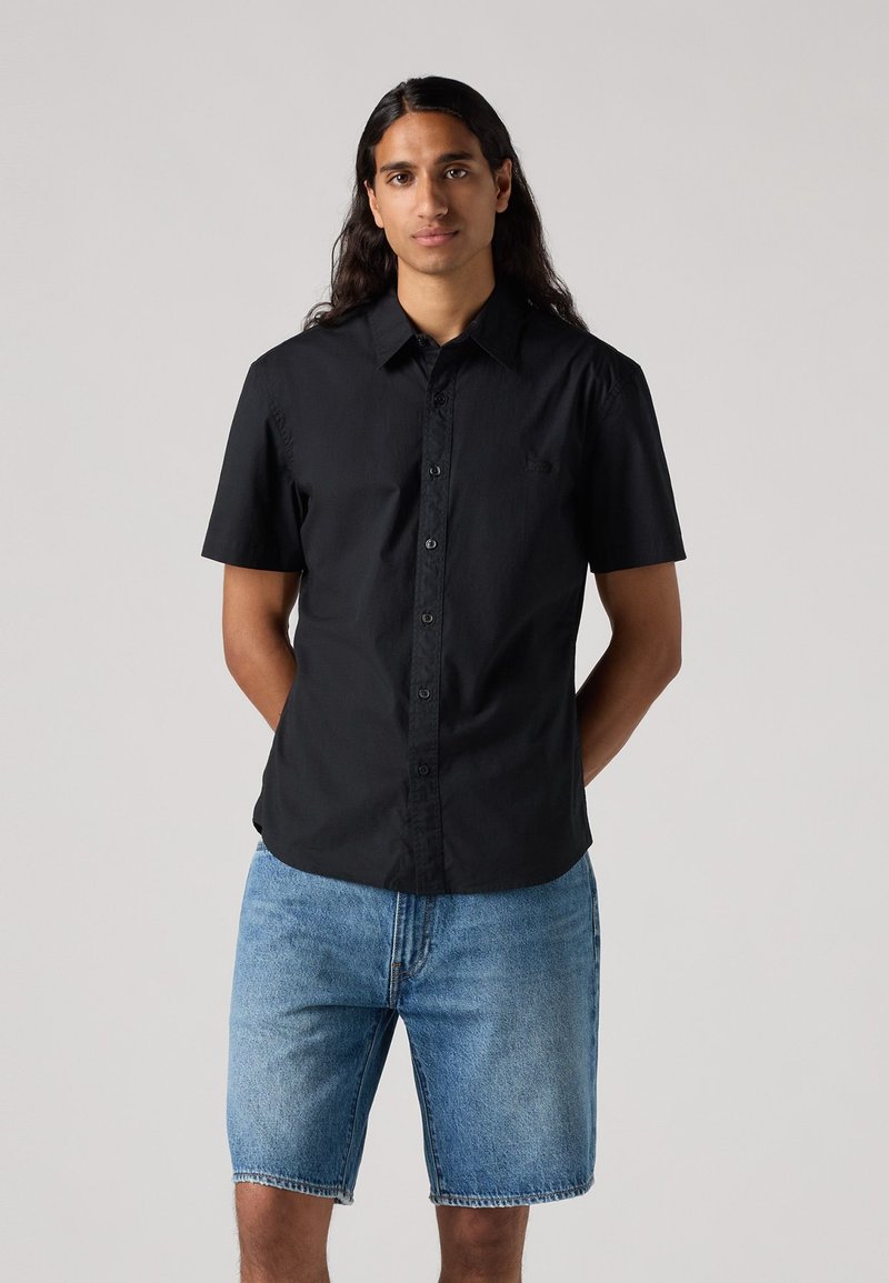 Maglietta nera a maniche corte con bottoni in tessuto leggero, abbinata a pantaloni corti in denim azzurro. Design semplice, senza motivi o accenti visibili.