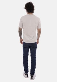 Uomo con capelli ricci, tatuaggi sulle braccia, indossa una maglietta bianca semplice, jeans blu scuro e scarpe da ginnastica bianche, in piedi di schiena.