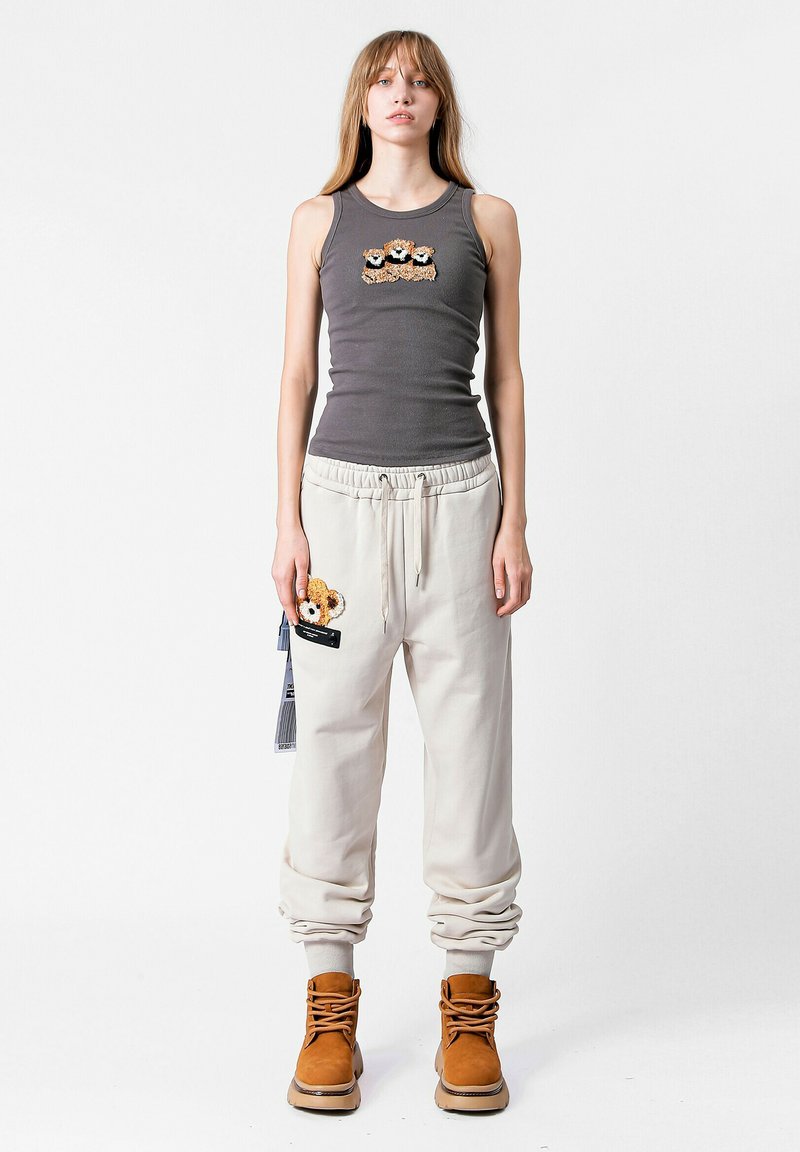 Camiseta sin mangas gris con un diseño de oso bordado, combinada con pantalones de chándal beige claros que tienen un parche de oso. Botas marrones con cordones completan el look.