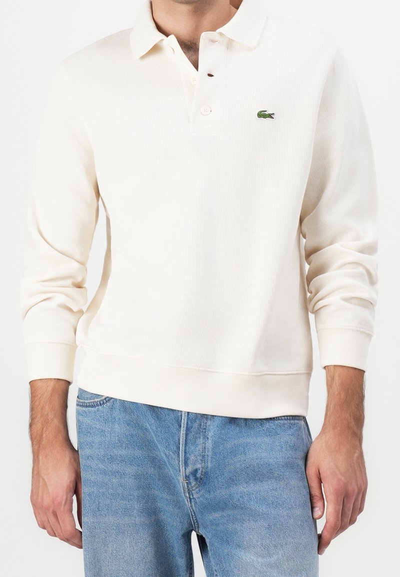 Homme portant un polo à manches longues couleur crème avec un logo crocodile vert et un jean bleu clair, debout sur fond blanc.