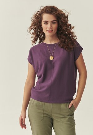 Göndör hajú nő lila rövid ujjú felsőt, olívazöld nadrágot és arany medálos nyakláncot visel, egyik kezével a zsebében áll.