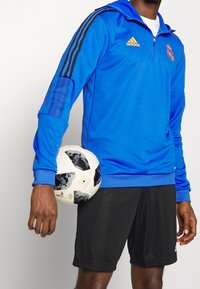 Personne portant une veste de sport bleue Adidas du Real Madrid et un short noir, tenant un ballon de football contre sa hanche.