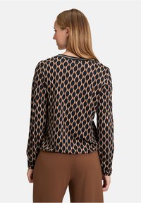 Maglietta nera a maniche lunghe con un motivo geometrico nei toni del beige e dell'arancione, scollatura rotonda e texture liscia, abbinata a pantaloni marroni.