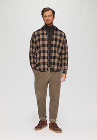 Chemise en flanelle à carreaux marron et noir, portée ouverte sur un turtleneck noir, assortie à un pantalon en velours côtelé beige et des baskets marron.