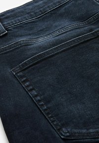 Tamno plave traperice s glatkom teksturom, ravnog kroja i stražnjim džepom s kontrastnim šavovima. Vidljivi su neuredni rubovi.