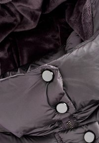 Interiorul jachetei tip puffer este de un purpuriu închis, având o textură moale de pluș, un material neted și butoane metalice cu detalii de fermoar.