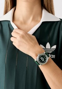 Montre de sport verte avec un cadran rond, des accents argentés, un bracelet vert texturé, et un haut de sport vert avec un col blanc et des rayures noires.