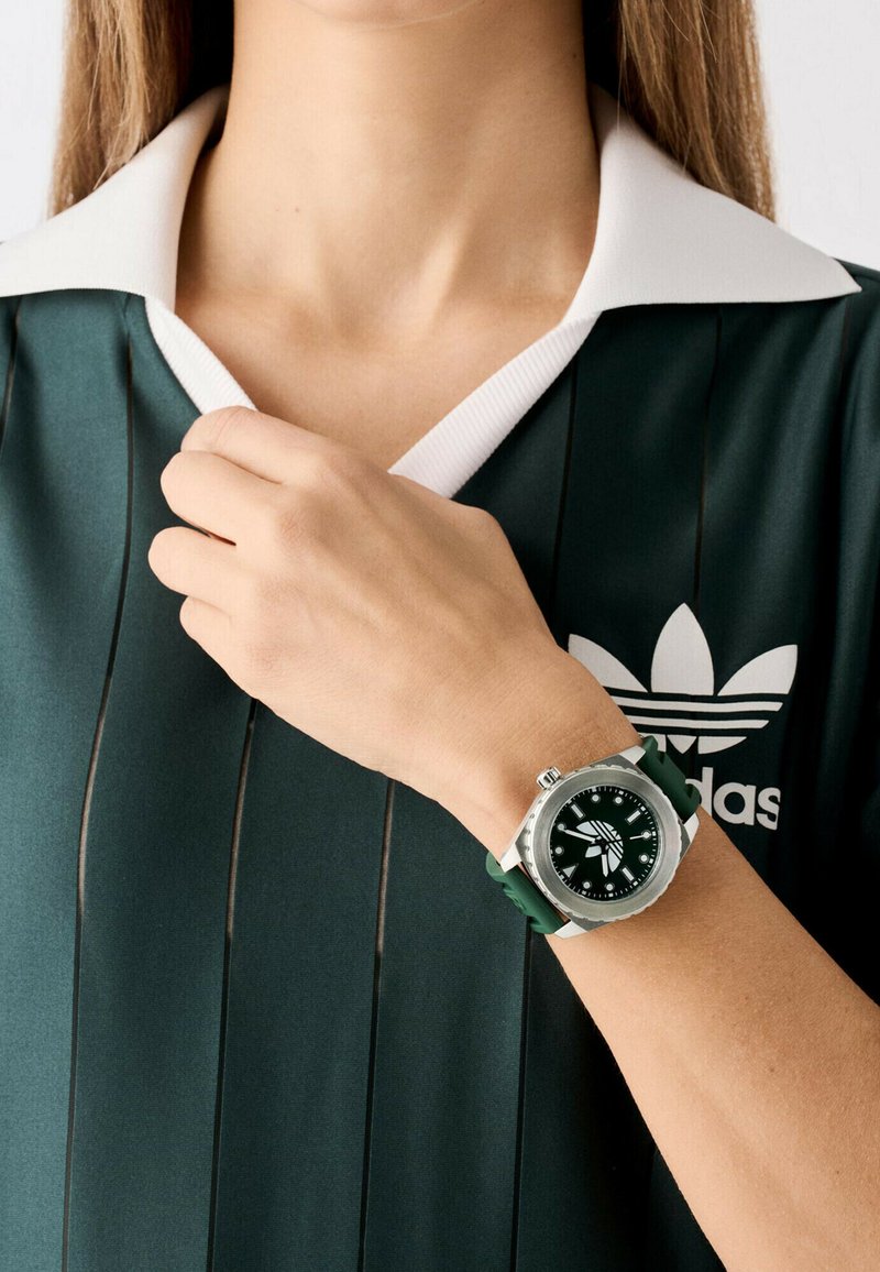 Montre de sport verte avec un cadran rond, des accents argentés, un bracelet vert texturé, et un haut de sport vert avec un col blanc et des rayures noires.