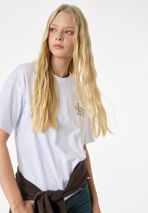 Ung kvinde med langt blond hår iført en hvid t-shirt med grøn tekst og en brun sweater bundet om livet.