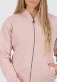 Giovane donna che indossa una giacca zip leggera di colore rosa chiaro con tasche, con una mano appoggiata nella tasca destra e capelli biondi lisci sulle spalle.